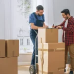 Customer signing a document while a professional mover looks on, holding a moving dolly with boxes.