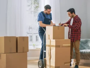 Customer signing a document while a professional mover looks on, holding a moving dolly with boxes.