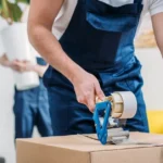 Professional mover taping a box shut while another mover holds a large house plant in the background